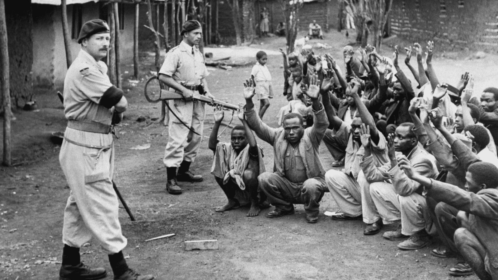A historical black-and-white photograph depicting a detention camp operation during the Mau Mau uprising in colonial-era Kenya. On the left, a British military or security officer wearing a beret and a light-colored uniform stands with his arms crossed, watching a large group of detained Kenyan men who are crouching on the dirt ground in rows with their hands raised. Behind the detainees, a second armed soldier or guard in uniform stands holding a rifle, while several women and children are visible in the background near simple, thatched-roof village or camp structures.