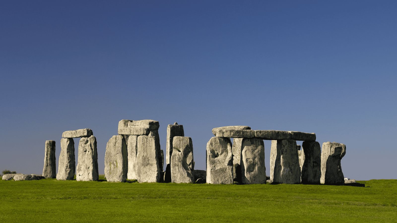 A wide, eye-level photograph of the prehistoric monument of Stonehenge standing on a vibrant green grass field in Wiltshire, England. The heavy sarsen stones and horizontal lintels form ancient, weathered archway structures under a vast, completely clear, and brilliant blue sky.