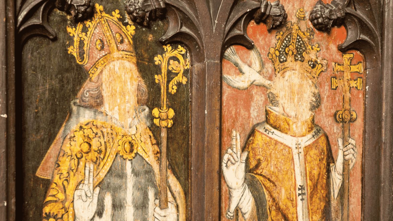 A close-up photograph of a medieval painted rood screen or wooden panel showing two religious figures whose faces have been intentionally defaced or scratched away, likely during the English Reformation. On the left, a bishop or archbishop wears a highly decorative golden mitre and cope, raising his right hand in blessing while holding an ornate golden crosier in his left. On the right, a crowned ecclesiastical figure or pope is depicted in golden and white vestments, raising two fingers in blessing and holding a papal ferula (triple cross staff), with a white dove, symbolizing the Holy Spirit, positioned near his shoulder against a faded red background.