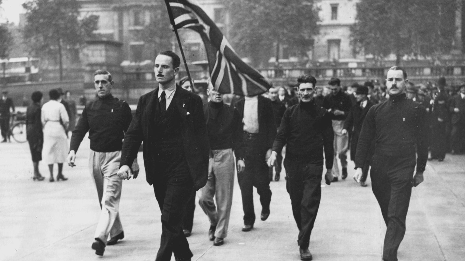 A historical black-and-white photograph showing Sir Oswald Mosley marching through a public street alongside members of the British Union of Fascists in the 1930s. Mosley walks prominently in the left foreground wearing a dark civilian suit, tie, and a small badge on his lapel. He is flanked by men in the movement's uniform, consisting of dark, high-necked blackshirts and trousers, with one man directly behind him carrying a large Union Jack flag that waves in the air as bystanders watch from the background.