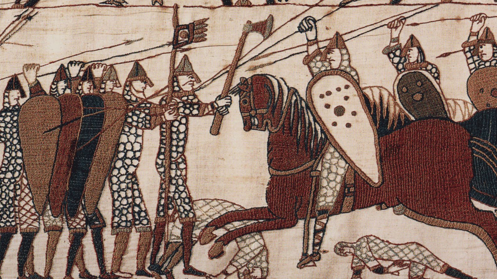 A close-up view of a section from the Bayeux Tapestry depicting a battle scene, likely from the Battle of Hastings. On the left, a disciplined shield wall of Anglo-Saxon housecarls and soldiers stands closely together, wearing patterned mail hauberks and pointed helmets with nasal guards, while one prominent warrior swings a large two-handed Dane axe against an enemy horse. On the right, Norman cavalrymen charge forward on horseback, brandishing spears and holding large, kite-shaped shields, with slain soldiers depicted lying on the ground along the lower border of the embroidered linen cloth.