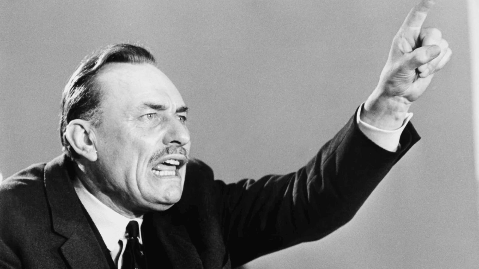A historical black-and-white photograph showing British politician Enoch Powell speaking passionately during a public address. Captured from the chest up, Powell is depicted mid-speech with an intense facial expression, looking toward the right with his mouth open and teeth bared. He wears a formal suit jacket, white collared shirt, and dark tie, while raising his left arm prominently across the frame, pointing his index finger upward against a plain, light background.