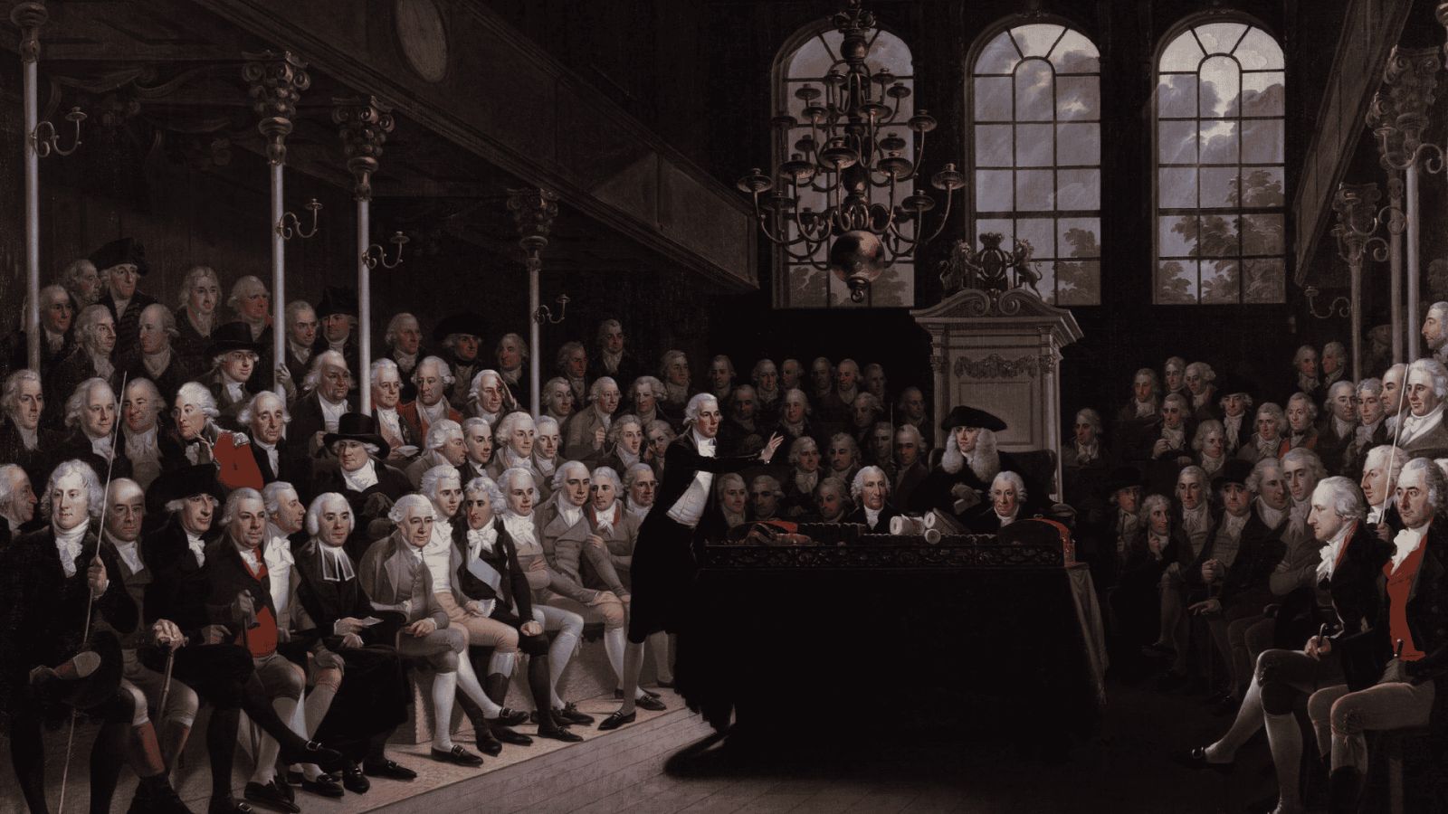 An 18th-century painting by Karl Anton Hickel depicting a session of the House of Commons in 1793. In the center foreground, William Pitt the Younger, the Prime Minister, stands addressing the chamber with his arm extended toward a large, dark wooden table strewn with documents and rolls of parchment. Behind the table, the Speaker of the House sits in an ornate, high-backed wooden chair beneath a large brass chandelier hanging from the ceiling. Densely packed rows of Members of Parliament, dressed in formal 18th-century coats, waistcoats, and powdered wigs, line the green benches on both sides of the chamber, listening attentively under high, arched windows.