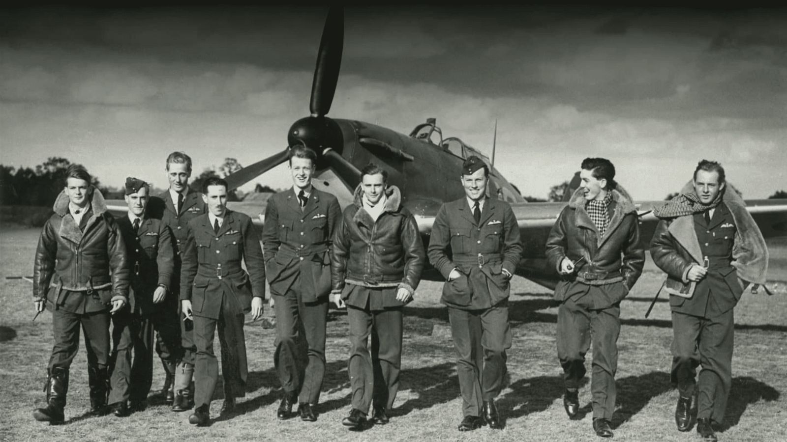 A historical black-and-white photograph showing a group of nine Royal Air Force (RAF) fighter pilots from No. 249 Squadron walking together across a grassy airfield during the Battle of Britain in September 1940. The pilots are dressed in standard RAF service dress uniforms, with several wearing heavy, fleece-lined leather flight jackets and silk scarves. They are walking side-by-side with relaxed, confident expressions beneath a cloudy sky, directly in front of a parked Hawker Hurricane fighter aircraft with its large propeller prominent in the background.